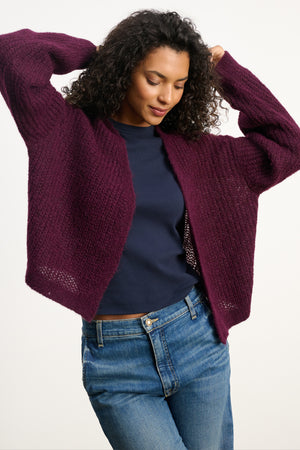 Wearing the Velvet by Graham & Spencer RANYA ALPACA CARDIGAN over a navy shirt and blue jeans, a person stands with arms raised, smiling and looking down, wrapped in this cozy essential.