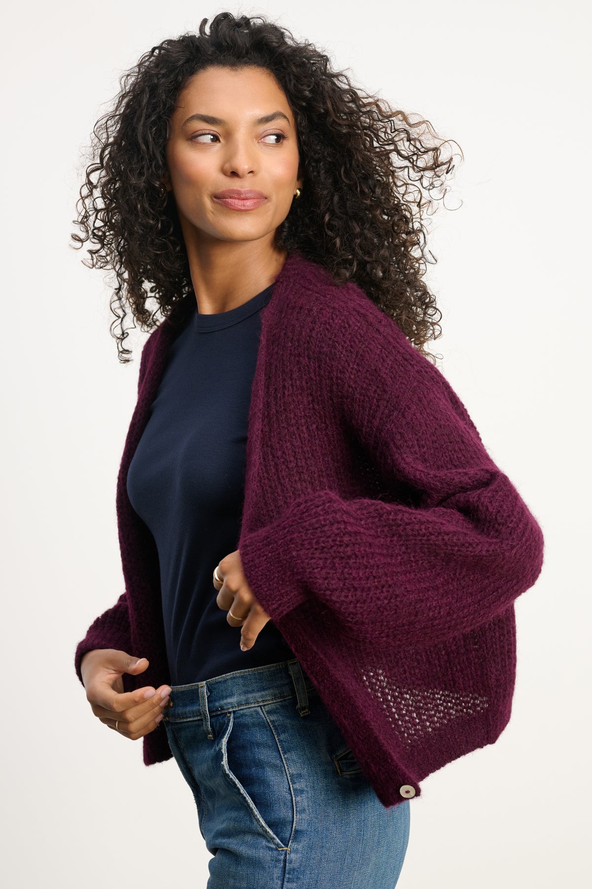 A woman with curly hair wears a dark blue shirt, blue jeans, and the maroon RANYA ALPACA CARDIGAN by Velvet by Graham & Spencer, standing against a plain light background.-40227142860993