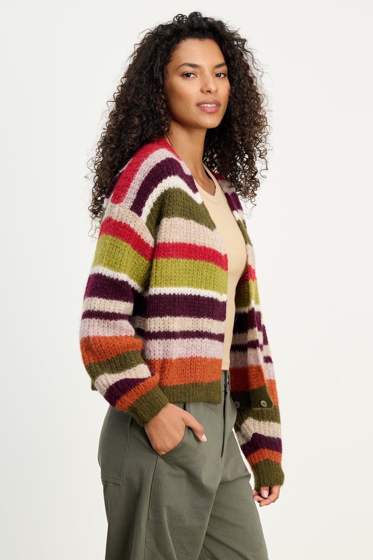   Woman with curly hair wears the RANYA ALPACA CARDIGAN by Velvet by Graham & Spencer—a cozy multicolored striped layer—over a beige top and olive green pants, standing against a plain light background. 