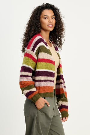Woman with curly hair wears the RANYA ALPACA CARDIGAN by Velvet by Graham & Spencer—a cozy multicolored striped layer—over a beige top and olive green pants, standing against a plain light background.