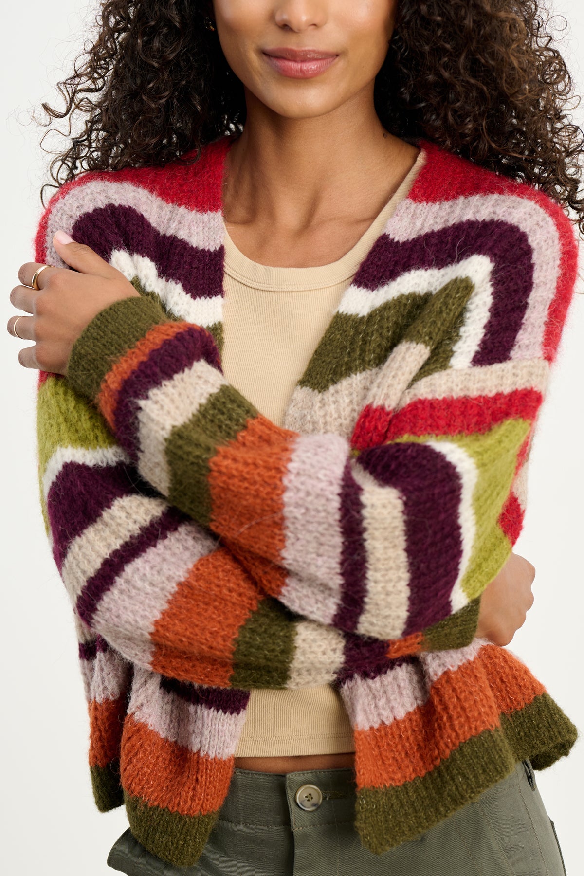 A woman wears the RANYA ALPACA CARDIGAN by Velvet by Graham & Spencer, a cozy wrap-style cardigan with textured, multicolored stripes in red, purple, green, orange, and beige over a tan top and olive green pants.-39742382735553