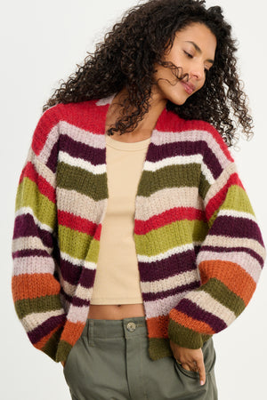 A woman with curly hair wears the RANYA ALPACA CARDIGAN by Velvet by Graham & Spencer, featuring colorful stripes, layered over a beige crop top and olive green pants, standing against a plain light background.