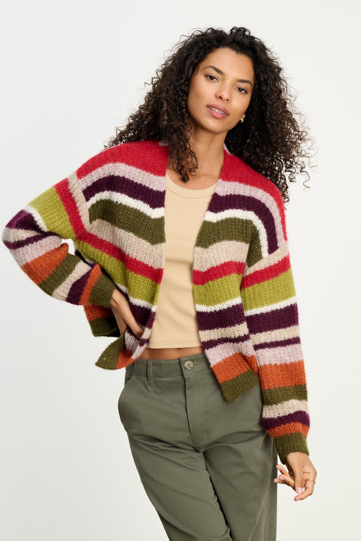 A woman with curly hair layers the Velvet by Graham & Spencer RANYA ALPACA CARDIGAN—multicolored and striped—over a beige top and olive green pants, standing against a plain light background.-39742382637249