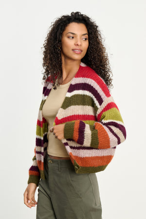 A woman with curly hair layers the Velvet by Graham & Spencer RANYA ALPACA CARDIGAN, featuring red, green, purple, and beige stripes, over a beige top and olive pants against a plain background.