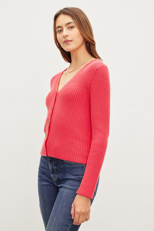 A woman in a red Velvet by Graham & Spencer HYDIE BUTTON FRONT CARDIGAN and blue jeans standing against a plain background, looking at the camera.
