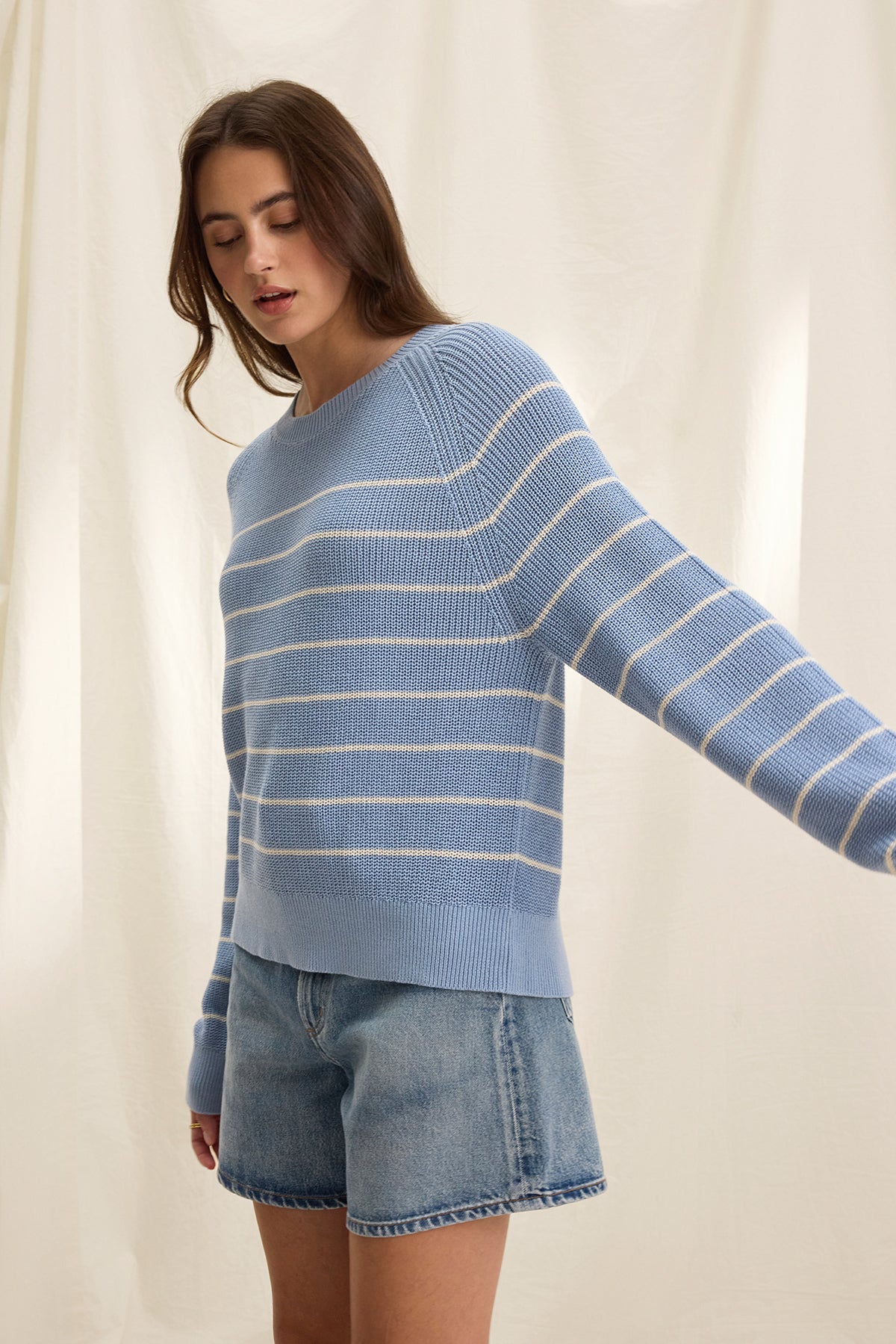   A person in a CHAYSE SWEATER by Velvet by Graham & Spencer, crafted from textured cotton, and denim shorts stands before a light-colored curtain, showcasing a relaxed silhouette ideal for casual wear. 