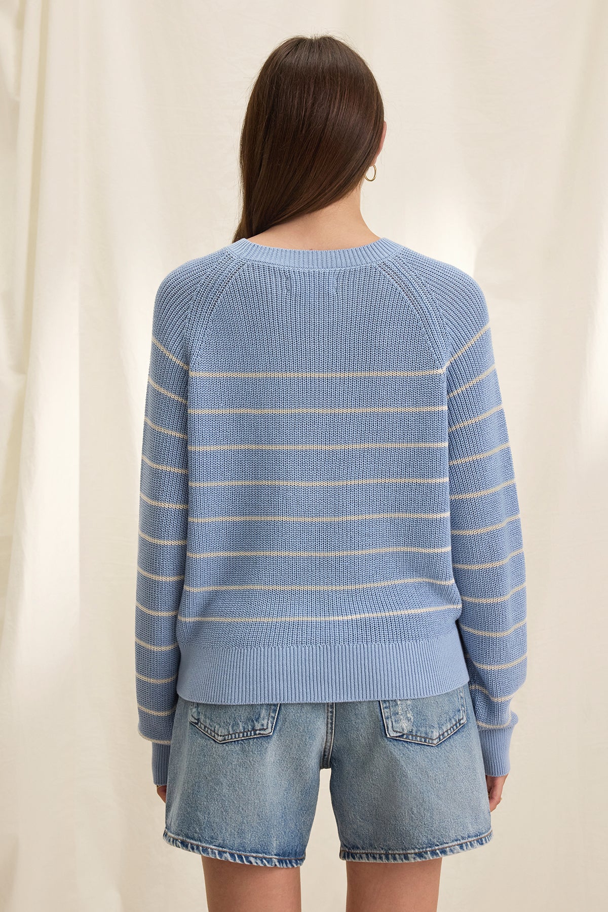   Back view of a person with long hair wearing the CHAYSE SWEATER by Velvet by Graham & Spencer, featuring a blue and white textured cotton stripe design and paired with denim shorts, creating a relaxed silhouette against a light background. 