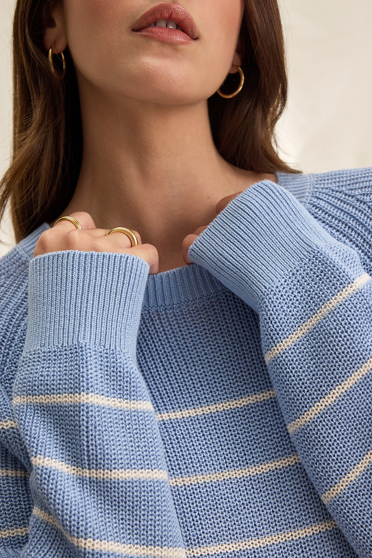   The individual is wearing the CHAYSE SWEATER from Velvet by Graham & Spencer, a light blue knit with white stripes and a relaxed fit. Their light brown hair highlights gold hoop earrings and rings. The textured cotton sweater's sleeves drape slightly over their hands, ideal for casual wear. 