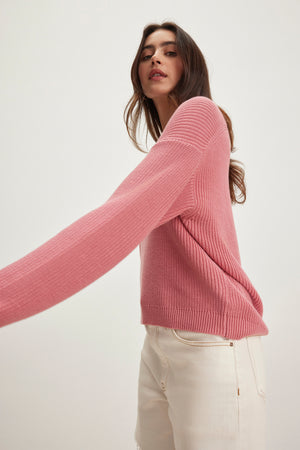 A person wearing the pink NEPAL SWEATER by Velvet by Graham & Spencer and white jeans stands against a plain light background, one arm extended outward.