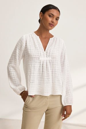 Wearing Velvet by Graham & Spencer's KORA Textured Cotton Gauze Top, a woman stands against a plain background in beige pants. With her right hand in her pocket, she confidently gazes at the camera.