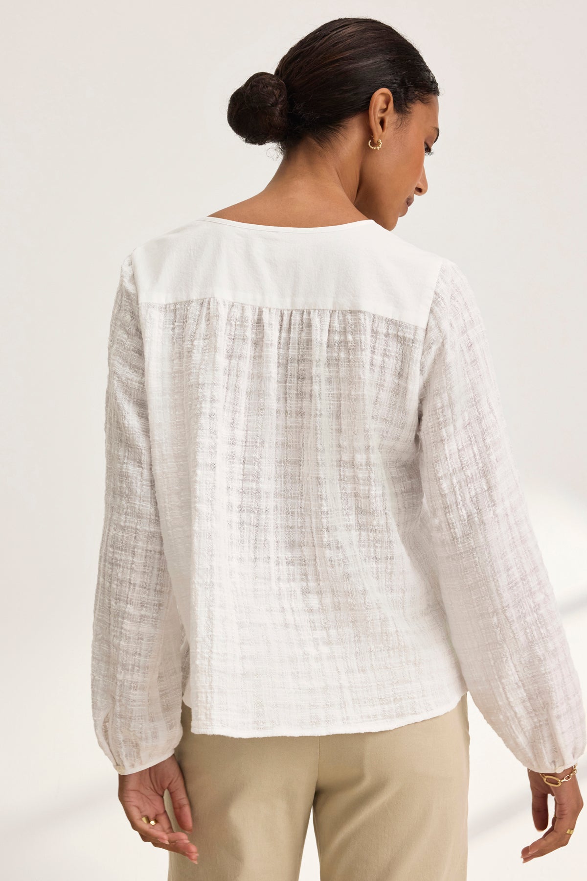 A woman in a KORA TEXTURED COTTON GAUZE TOP by Velvet by Graham & Spencer and beige pants stands gracefully turned away, her hair styled in a bun.-38943266930881