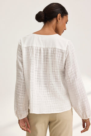 A woman in a KORA TEXTURED COTTON GAUZE TOP by Velvet by Graham & Spencer and beige pants stands gracefully turned away, her hair styled in a bun.