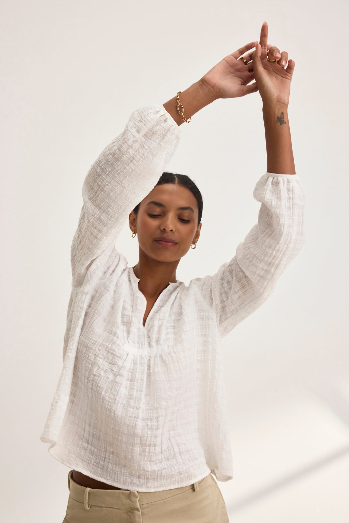 A person in a white KORA TEXTURED COTTON GAUZE TOP by Velvet by Graham & Spencer and beige pants, hands raised, eyes closed, showing a small tattoo on the right forearm.-38943266996417