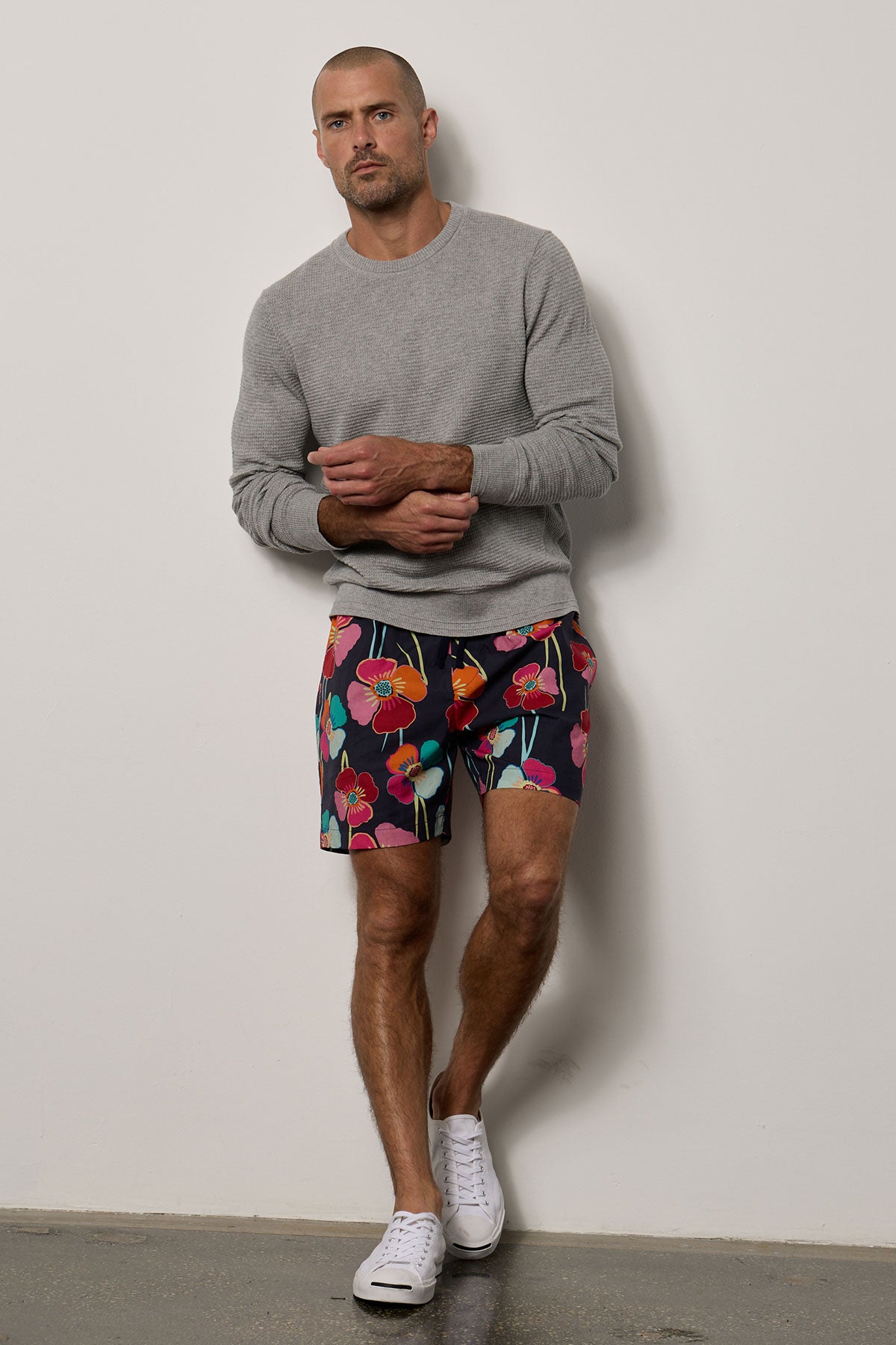 A man in Velvet by Graham & Spencer shorts and a grey sweatshirt is leaning against a wall, giving off a casual vacation look.-35678353490113