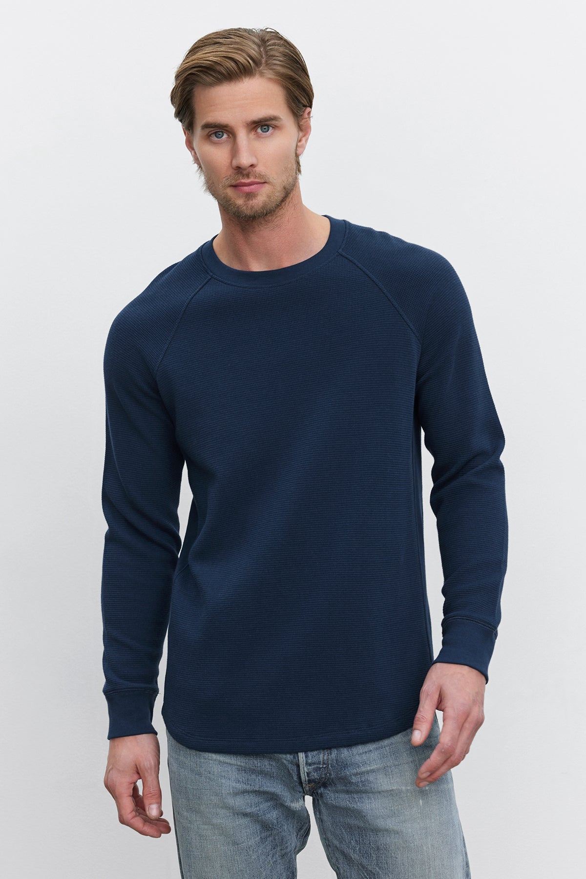   A man with short hair and a beard is wearing the GLEN THERMAL CREW by Velvet by Graham & Spencer, a navy blue long-sleeved cotton thermal shirt with a classic neckline, paired with blue jeans. He stands against a plain white background. 