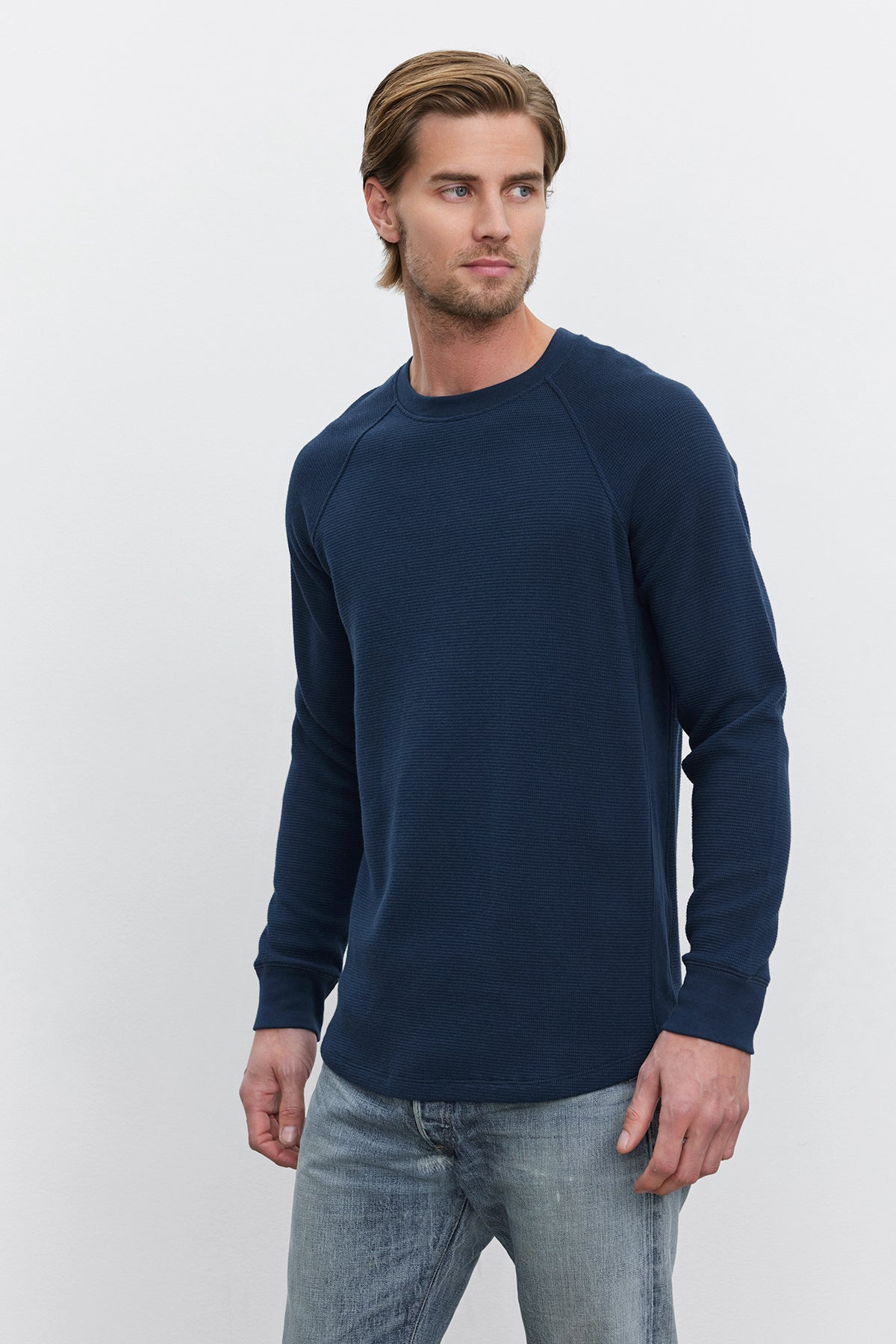   A man with light brown hair is wearing the GLEN THERMAL CREW by Velvet by Graham & Spencer in navy blue, paired with light blue jeans, and standing against a plain white background. 