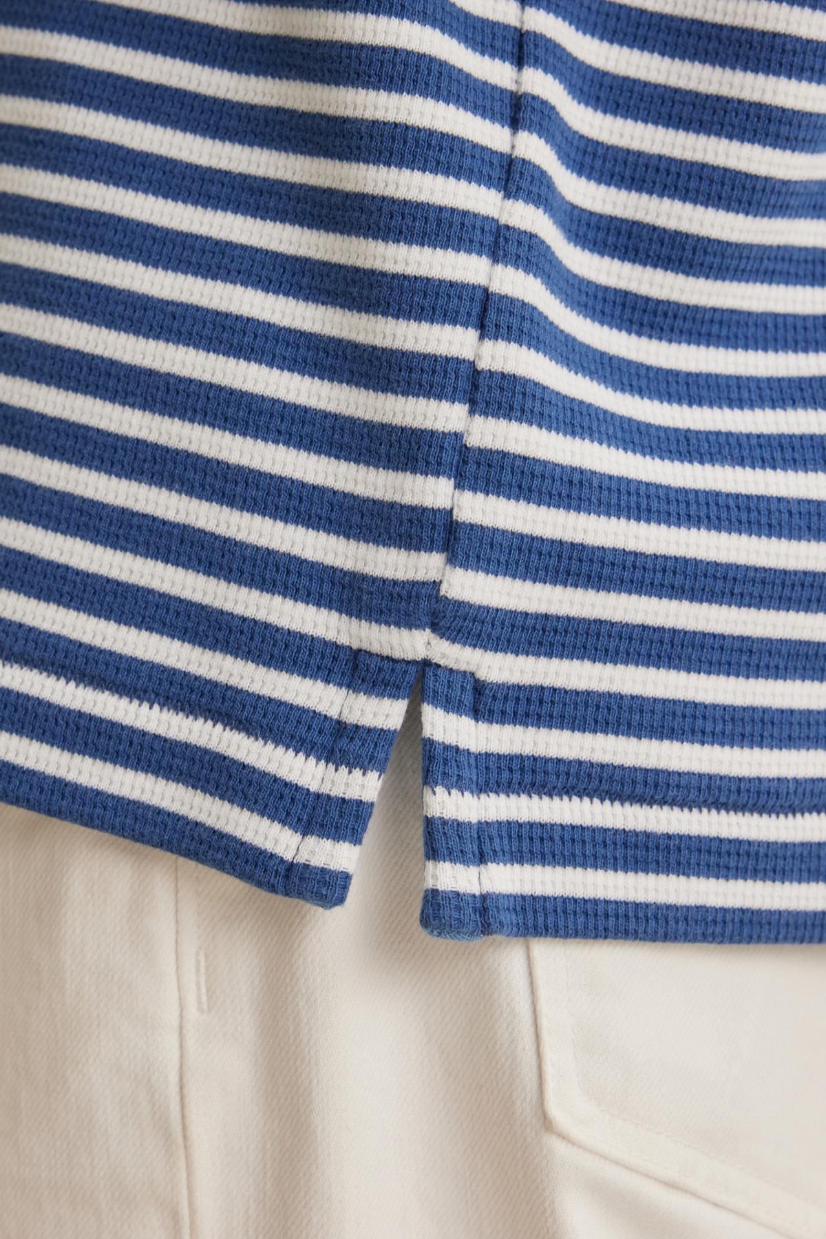   Close-up of the HARLAN THERMAL STRIPED CREW by Velvet by Graham & Spencer, featuring blue and white stripes with a small side split, paired with white pants. 