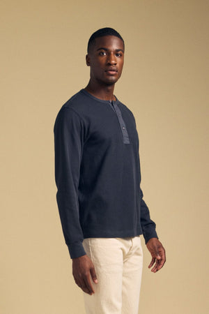 Front view of a man wearing the graphite CHANCE THERMAL HENLEY showcasing its button placket and long sleeves with a beige background