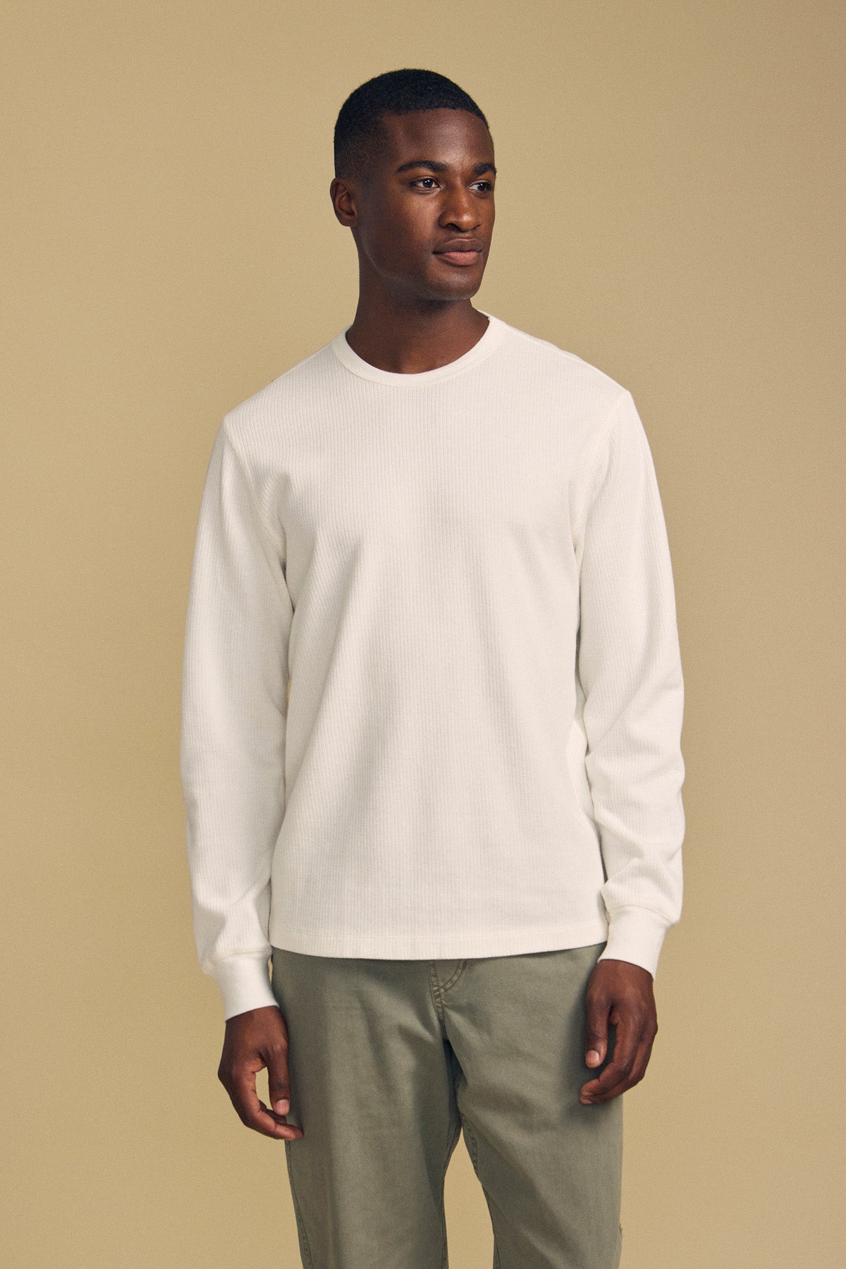 A man stands facing slightly left, wearing the Velvet by Graham & Spencer DARIEL THERMAL PULLOVER in white and light olive pants, against a plain beige background.