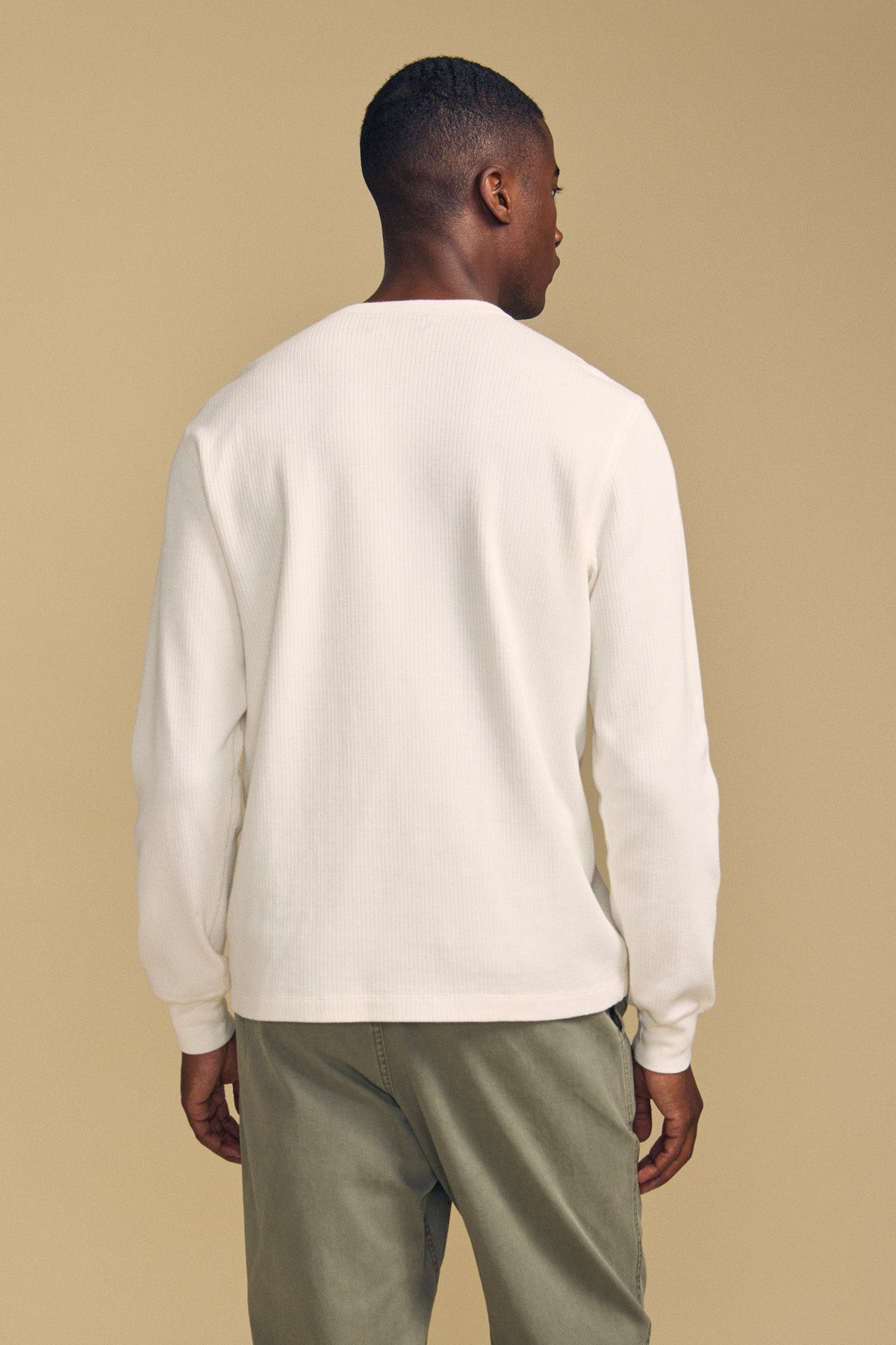A man stands facing away from the camera, wearing the Velvet by Graham & Spencer DARIEL THERMAL PULLOVER—a long-sleeve white shirt—paired with olive green pants against a plain beige background.-41859631087809