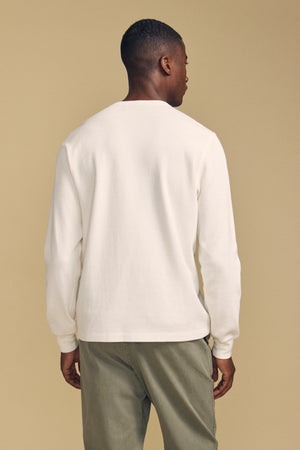 A man stands facing away from the camera, wearing the Velvet by Graham & Spencer DARIEL THERMAL PULLOVER—a long-sleeve white shirt—paired with olive green pants against a plain beige background.