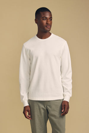 A man stands facing slightly left, wearing the Velvet by Graham & Spencer DARIEL THERMAL PULLOVER in white and light olive pants, against a plain beige background.