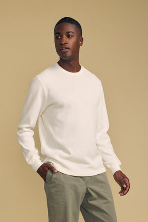 A man stands against a beige background wearing the DARIEL THERMAL PULLOVER by Velvet by Graham & Spencer—a white long-sleeve shirt—paired with light olive-green pants, with one hand in his pocket.