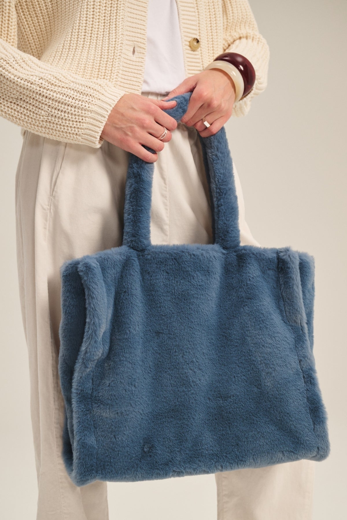 A person carries the LIBBY LUXE FAUX FUR TOTE by Velvet by Graham & Spencer, a stylish blue accessory, paired with light pants, a white top, and a beige knit cardigan—perfect for colder weather.-40268358058177