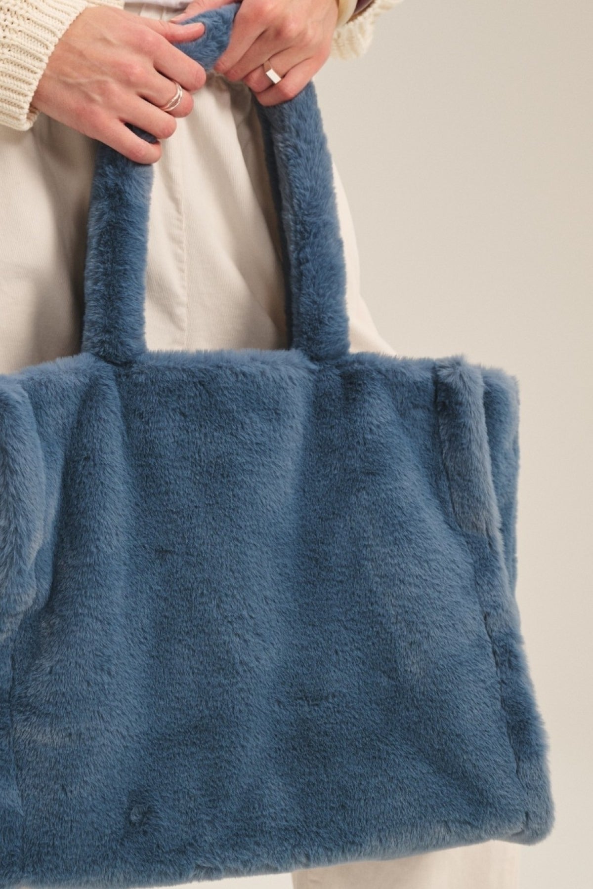 A person holds the LIBBY LUXE FAUX FUR TOTE by Velvet by Graham & Spencer, a large rectangular blue bag with two handles—an ideal cold-weather accessory.