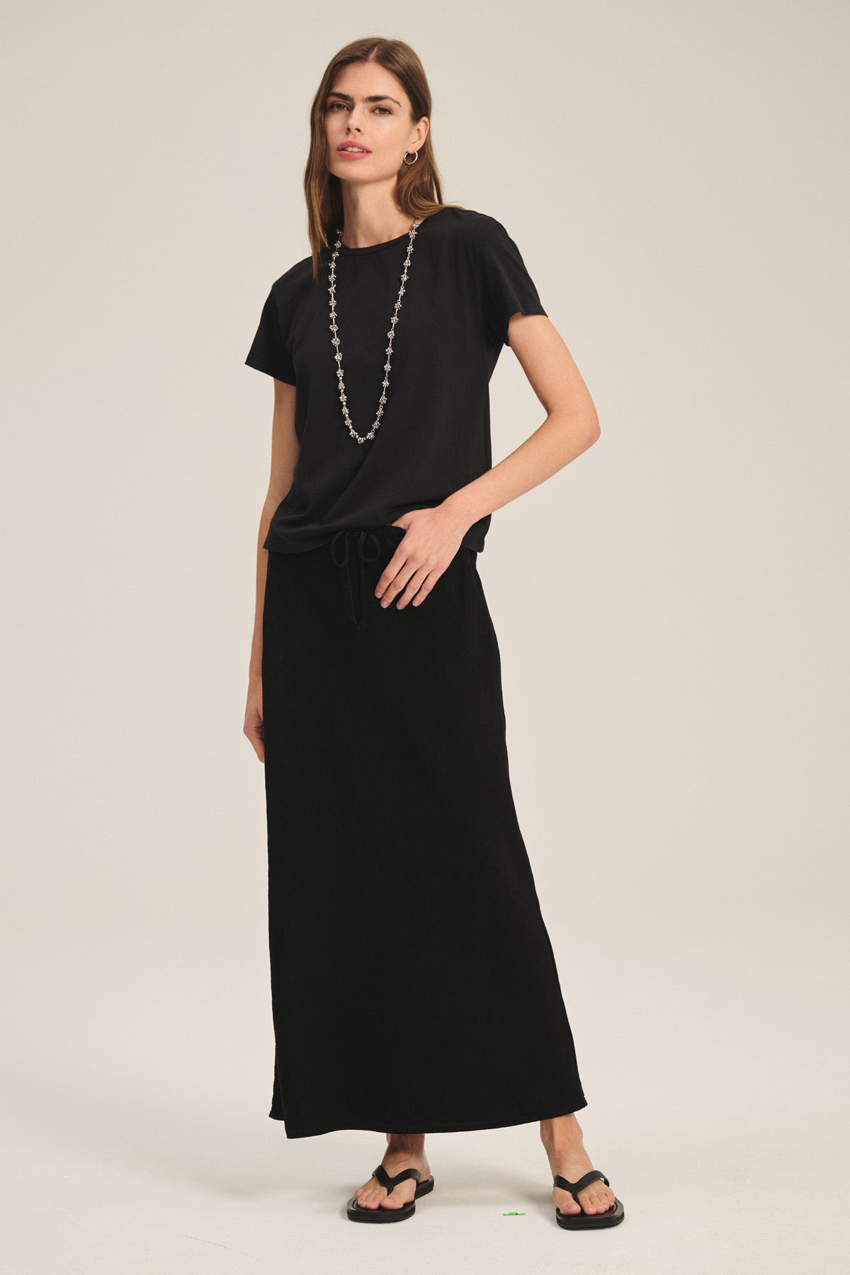 A woman wears the Velvet by Graham & Spencer TIA TEE, a classic black cotton jersey crewneck, styled with a long black skirt, flip-flops, and a beaded necklace against a plain background.