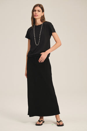 A woman wears the Velvet by Graham & Spencer TIA TEE, a classic black cotton jersey crewneck, styled with a long black skirt, flip-flops, and a beaded necklace against a plain background.