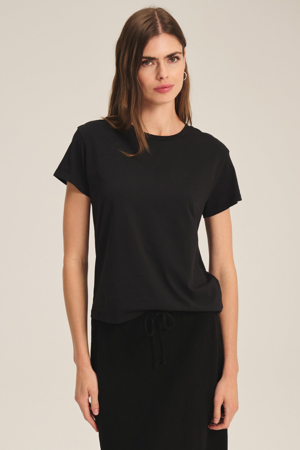 A woman with long brown hair wears the TIA TEE by Velvet by Graham & Spencer—a black cotton jersey crewneck—paired with a black skirt, standing against a plain light background.-40305565597889