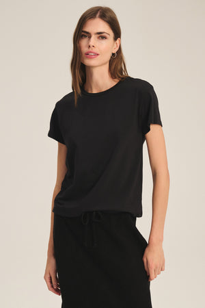 A woman stands against a plain background, wearing the TIA TEE by Velvet by Graham & Spencer—a classic black cotton jersey crewneck tee—paired with a black skirt, her left hand by her side.