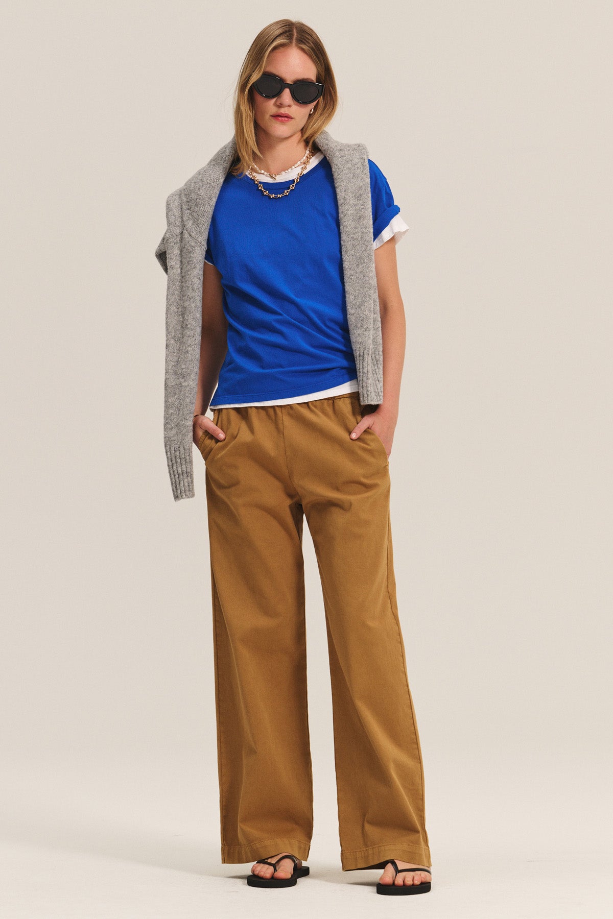 A person in a blue T-shirt over a white shirt, black sandals, sunglasses, and a gray sweater draped over their shoulders stands with hands in pockets, wearing the NAYA PANT by Velvet by Graham & Spencer in tan stretch cotton twill.-40672442974401