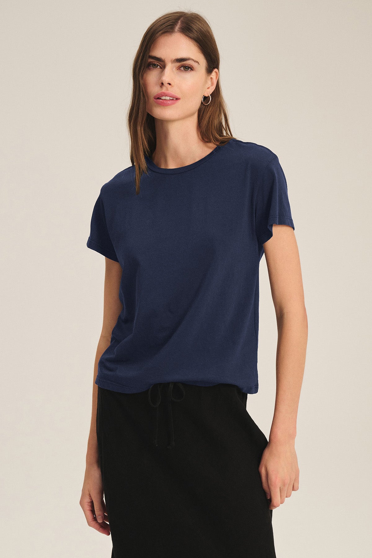A woman stands against a plain background wearing the Velvet by Graham & Spencer TIA TEE in navy blue cotton jersey and a black skirt, with one hand by her side—a timeless essential for any wardrobe.