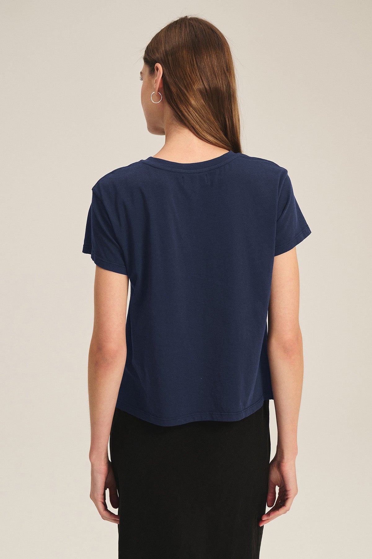 A person with long brown hair is seen from the back wearing the Velvet by Graham & Spencer TIA TEE in navy cotton jersey with a black skirt—an essential, timeless look against a plain background.