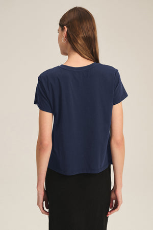 A person with long brown hair is seen from the back wearing the Velvet by Graham & Spencer TIA TEE in navy cotton jersey with a black skirt—an essential, timeless look against a plain background.