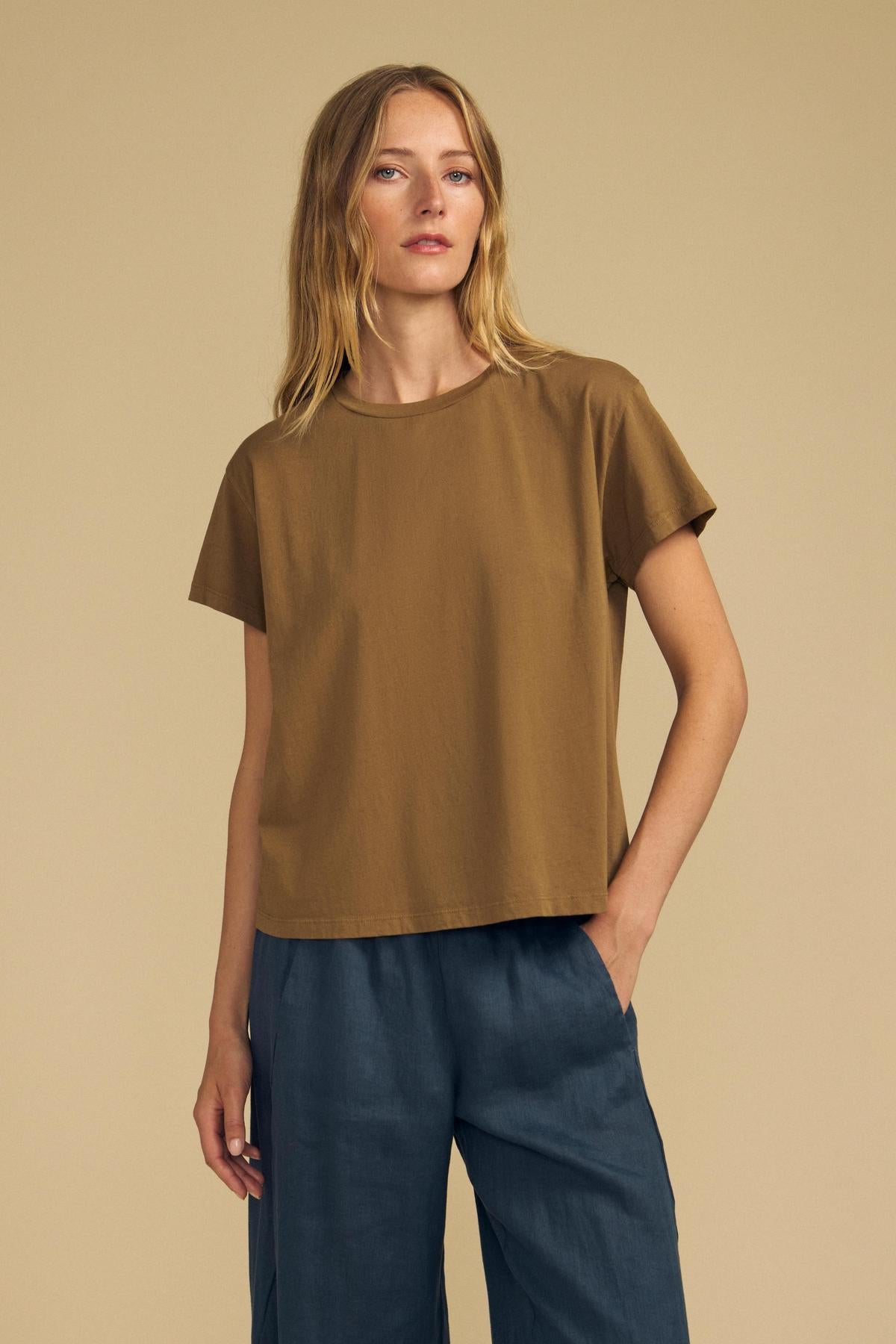 A person with long blonde hair wears the TIA TEE by Velvet by Graham & Spencer—a short-sleeve brown cotton jersey crewneck—paired with loose dark blue pants, standing against a beige background.