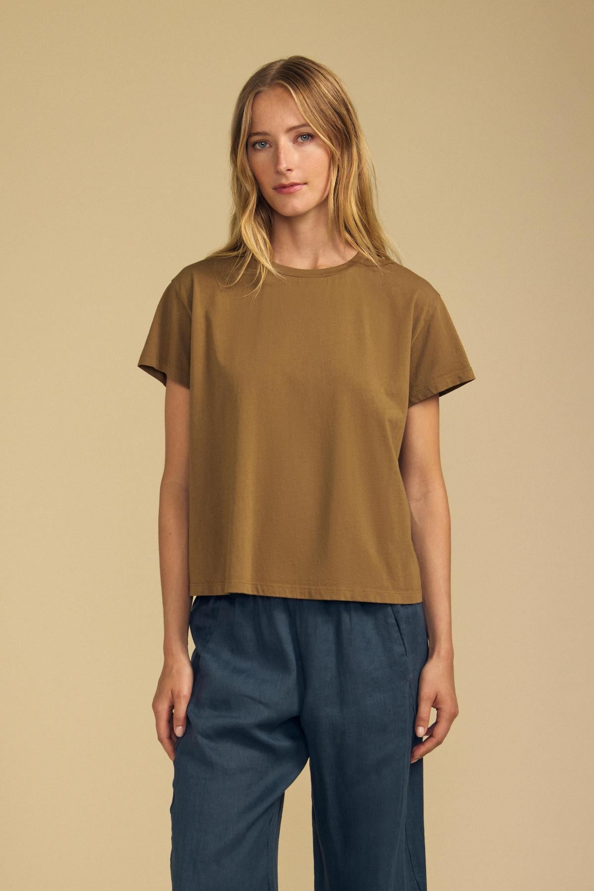 A woman stands against a beige background wearing the TIA TEE by Velvet by Graham & Spencer—a brown cotton jersey crewneck—and blue wide-leg pants, looking at the camera with a neutral expression.