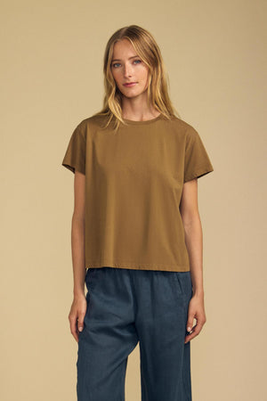 A woman stands against a beige background wearing the TIA TEE by Velvet by Graham & Spencer—a brown cotton jersey crewneck—and blue wide-leg pants, looking at the camera with a neutral expression.