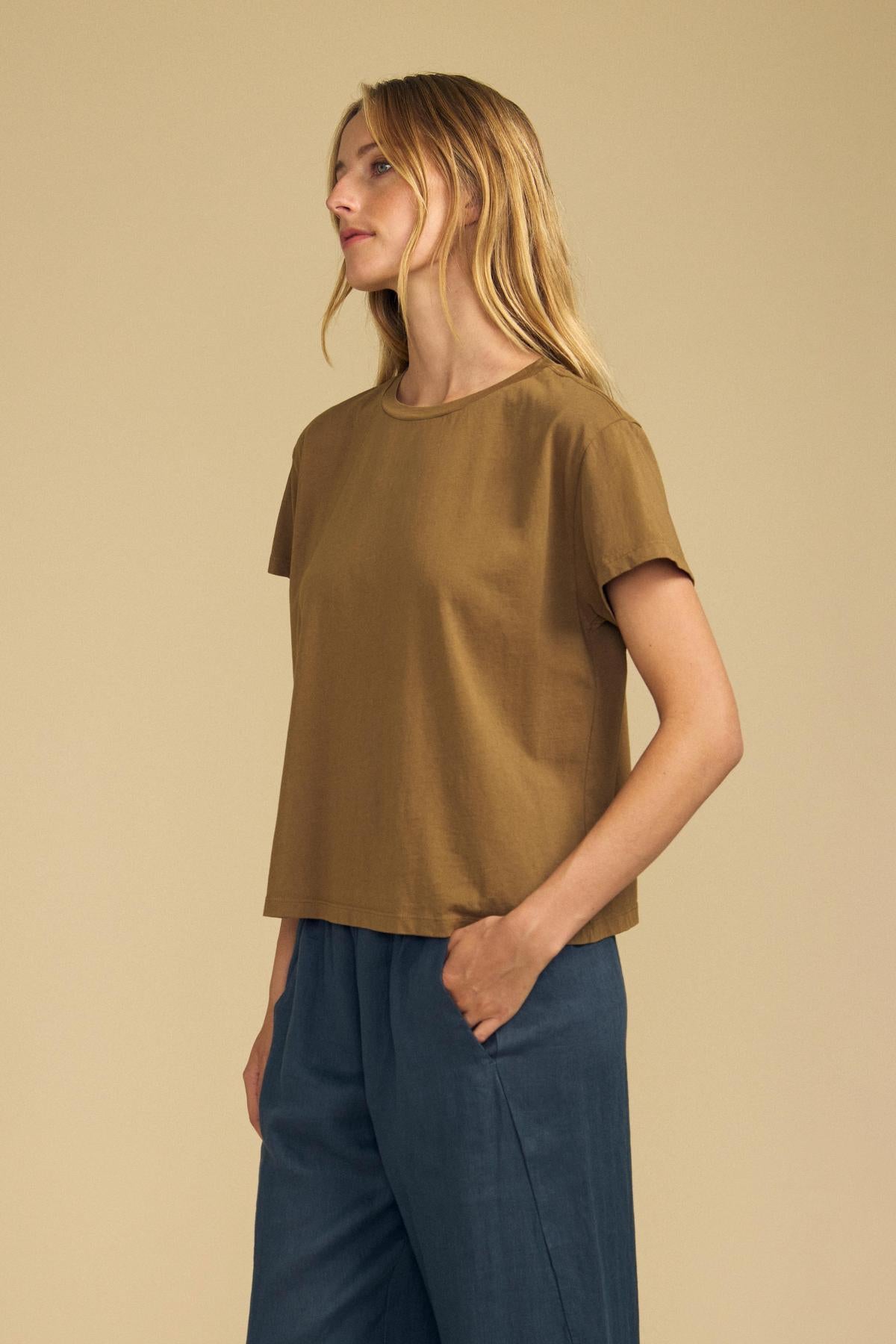 Woman stands against a beige background, wearing the Velvet by Graham & Spencer TIA TEE—a timeless essential brown cotton jersey crewneck—and blue pants, with her left hand in her pocket, looking to her left.