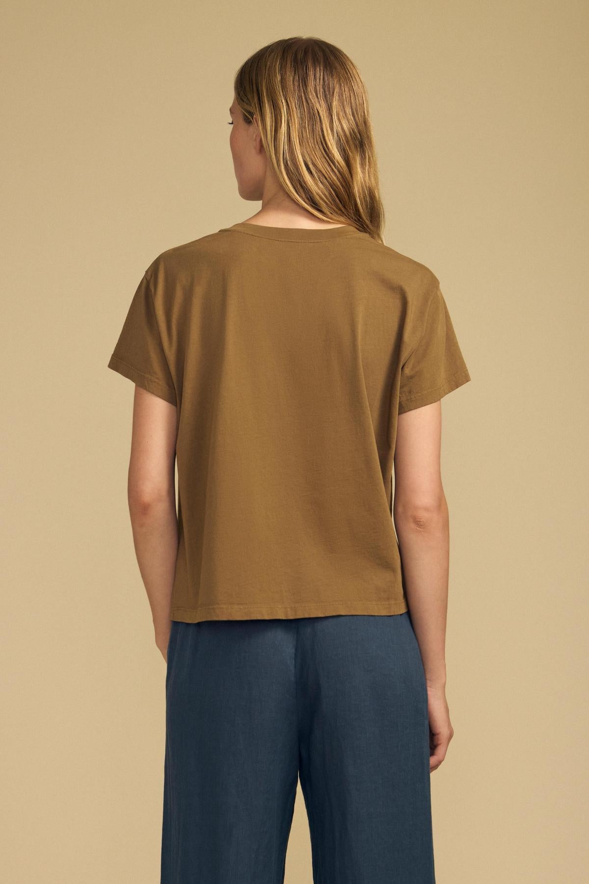 A person stands with their back to the camera, wearing the Velvet by Graham & Spencer TIA TEE in brown cotton jersey and blue pants against a plain beige background—a timeless essential for any wardrobe.