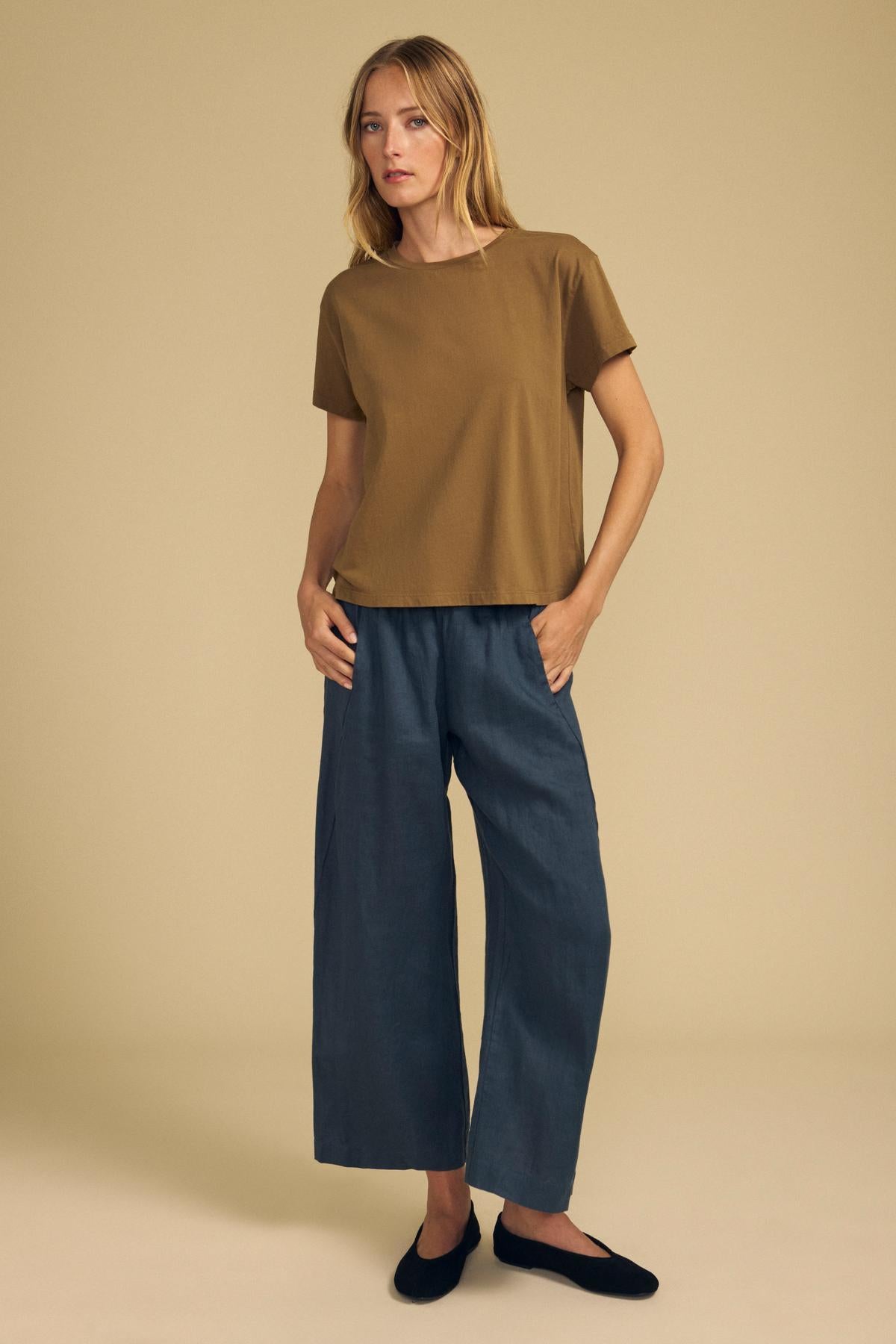 A woman stands against a beige background wearing the TIA TEE by Velvet by Graham & Spencer—a timeless crewneck crafted from soft cotton jersey—paired with wide-leg blue pants and black flats for an effortlessly chic look.-41996879429825
