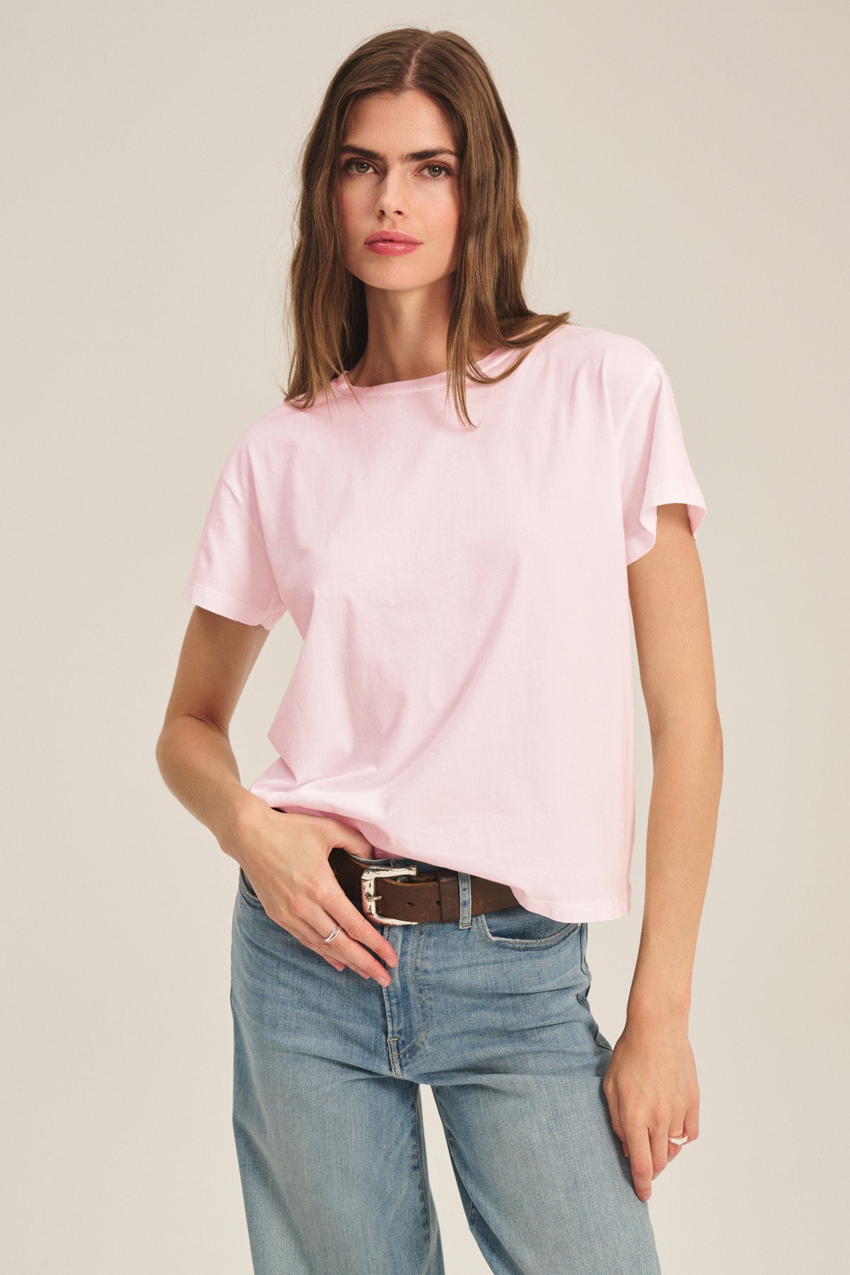 A woman stands against a neutral background wearing the TIA TEE by Velvet by Graham & Spencer—a light pink cotton jersey crewneck paired with blue jeans and a brown belt, creating a timeless wardrobe essential.-41044148453569