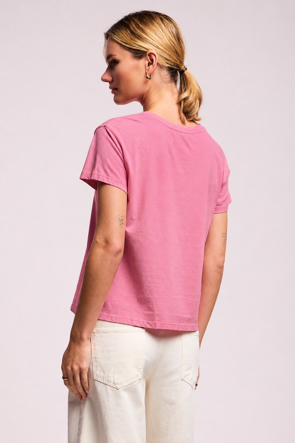 A woman stands with her back turned, wearing the TIA TEE by Velvet by Graham & Spencer in pink cotton jersey and off-white pants, set against a plain light background.-41393532403905