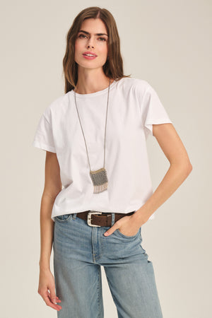 A woman wears the TIA TEE by Velvet by Graham & Spencer—a classic white crewneck cotton jersey tee—paired with blue jeans, a brown belt, and a long pendant necklace, standing against a neutral background.