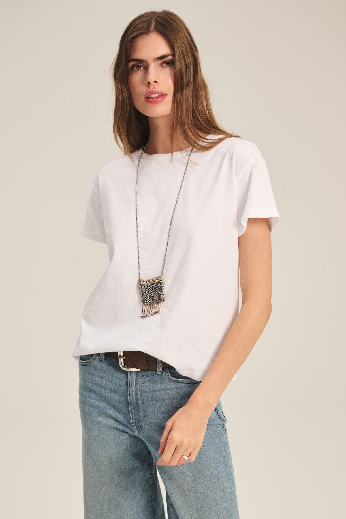 A woman wears the TIA TEE by Velvet by Graham & Spencer—a classic white cotton jersey crewneck—paired with blue jeans and a rectangular pendant necklace, standing against a neutral background.