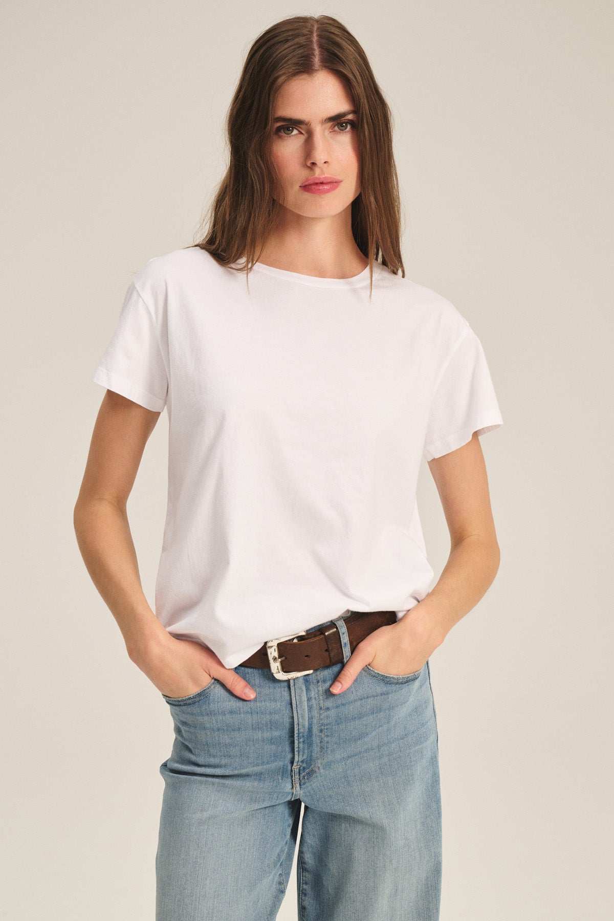 A woman with long brown hair wears the TIA TEE by Velvet by Graham & Spencer—a classic white cotton jersey crewneck tee—paired with blue jeans and a brown belt, standing hands-in-pockets against a neutral background.-40305566056641