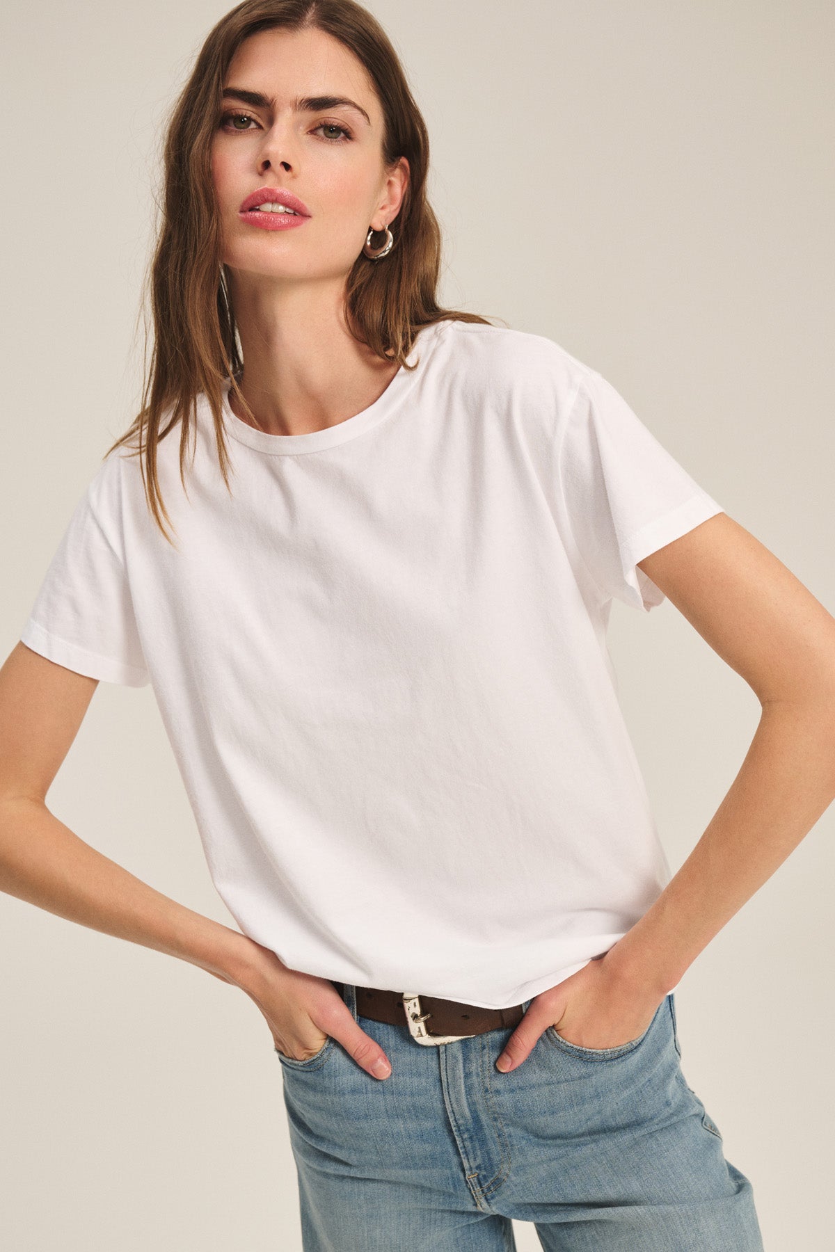 Someone wearing the Velvet by Graham & Spencer TIA TEE—a classic white crewneck made from soft cotton jersey—paired with light blue jeans stands with hands on hips against a neutral background.-40305565925569