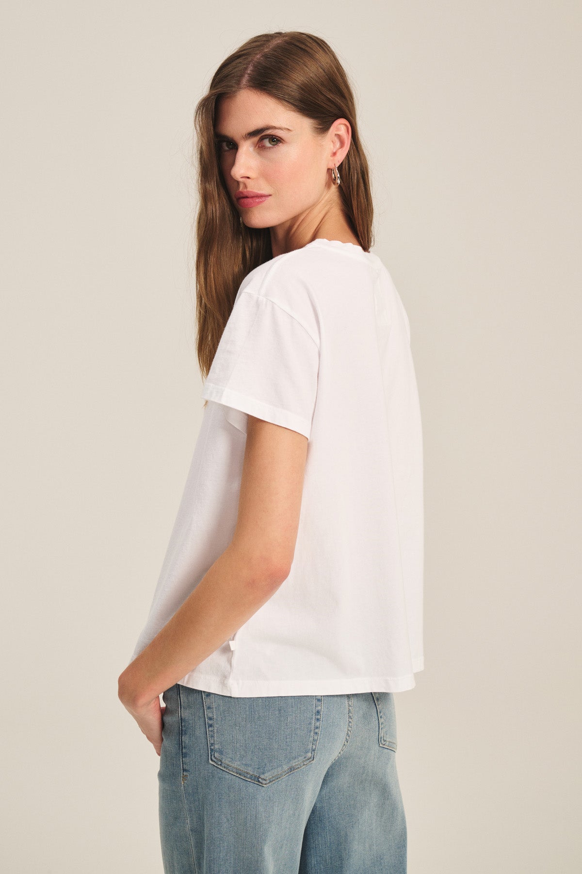A woman in the TIA TEE by Velvet by Graham & Spencer and blue jeans stands sideways, looking over her shoulder against a plain background—a classic staple for effortless style.-40305565761729